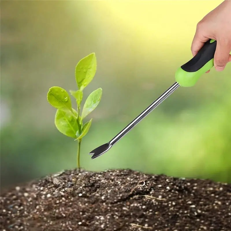 A hand holds a green and black stainless steel weeder tool, poised above a small green plant growing in dark soil.