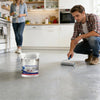 A man applies self-leveling epoxy floor paint to a concrete floor in a modern kitchen setting.