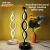 A black and white wavy LED desk lamp illuminates a bouquet of yellow and blue flowers on a wooden surface.
