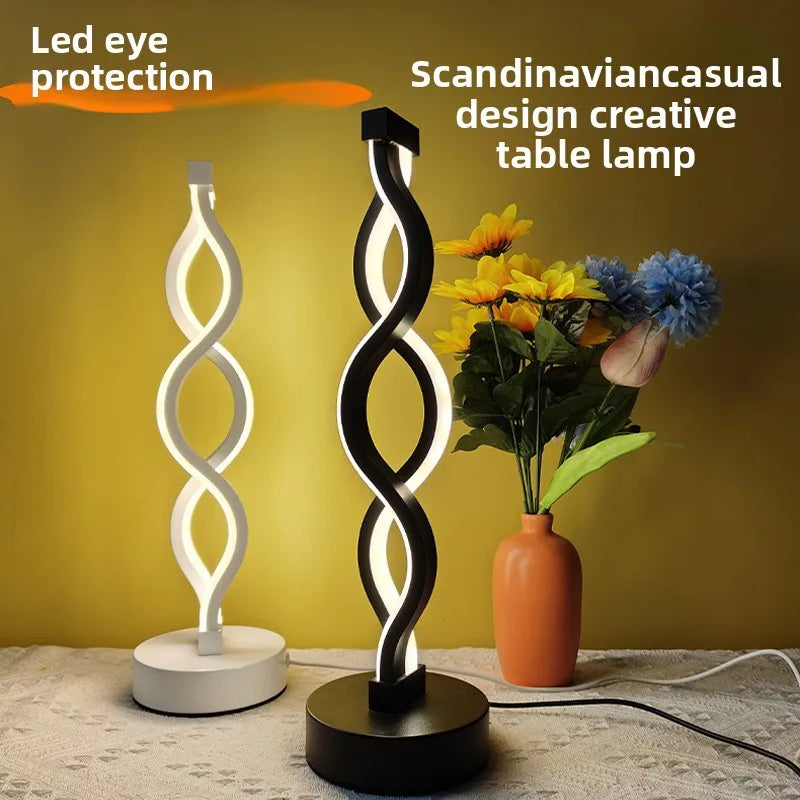 A black and white wavy LED desk lamp illuminates a bouquet of yellow and blue flowers on a wooden surface.