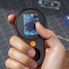 A hand holds a compact digital handheld microscope with an LED light, showing a circuit board under magnification.