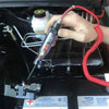 A hand holds a digital automotive voltage tester probe connected to a red cable, positioned near an engine component.