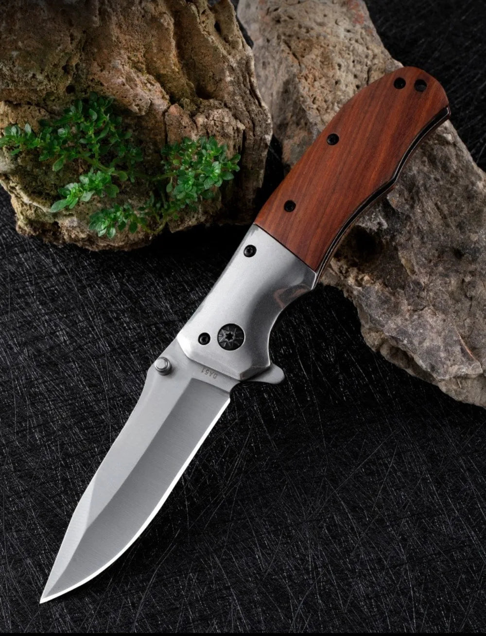 A premium folding utility pocket knife with a wooden handle and silver blade resting on dark stone next to green moss.