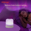 A white touch night light lamp emitting a purple glow is placed on a table next to a person resting in bed.