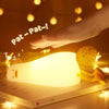 A hand gently pats a glowing silicone night light, illuminating a surface in a warm, soft light.