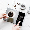 Hands are using a digital smart kitchen scale to measure coffee beans on a marble surface.
