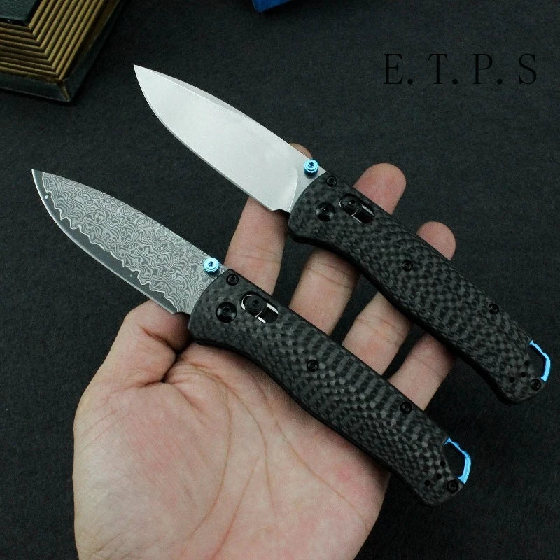 A hand holds a black Damascus steel folding hunting knife with a silver blade and blue accents.