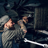 A bearded man in a cap is using a wrench to work on a tire in a dark garage setting.
