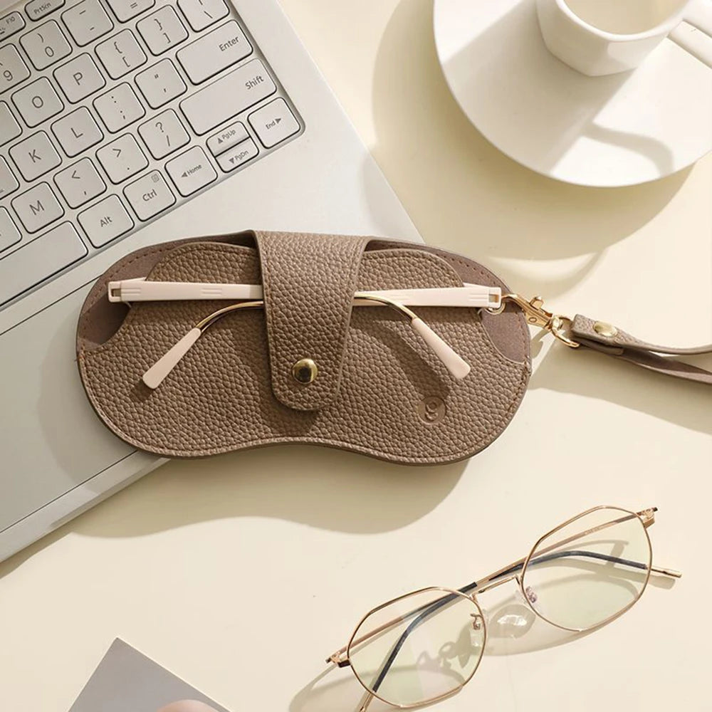 A tan PU leather eyeglasses case with a gold chain is shown next to a laptop keyboard and a pair of clear-framed glasses.