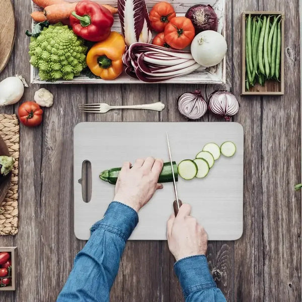 Hände schneiden eine Gurke auf einem Edelstahl-Cuttingboard umgeben von frischem Gemüse.
