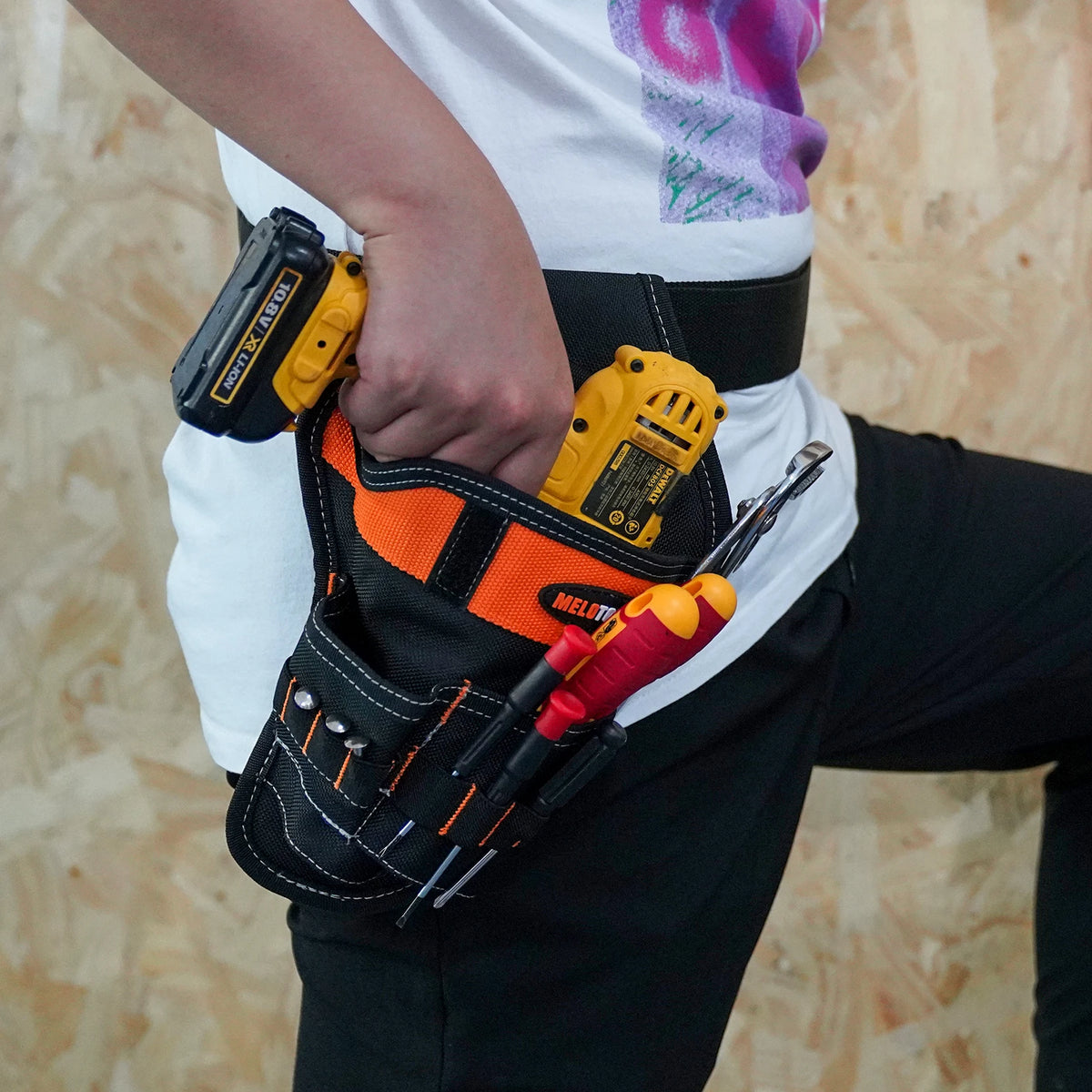 A hand holds a black tool holster containing yellow cordless drill bits and a red tool.