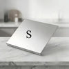 A square stainless steel cutting board with a single embossed letter S resting on a white marble surface.