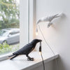 Eine schwarze Vogelskulptur hängt an einer weißen Wand neben einer modernen Tischleuchte.