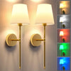 A pair of modern brass wall sconces with white lampshades and color-changing light options mounted on a textured wall.