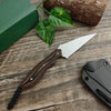 A compact fixed blade utility knife with a wooden handle and silver blade resting on a rustic wooden surface next to a green 