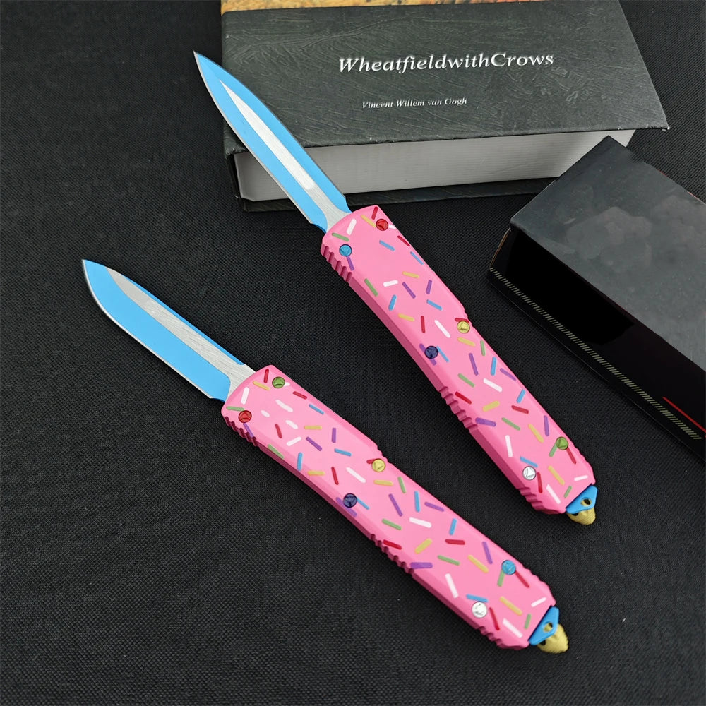 Pink folding knife with a D2 blade and aluminum handle resting on a dark surface next to a box.