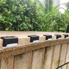A row of solar powered deck and pathway lights mounted on a textured stone wall surrounded by lush greenery.