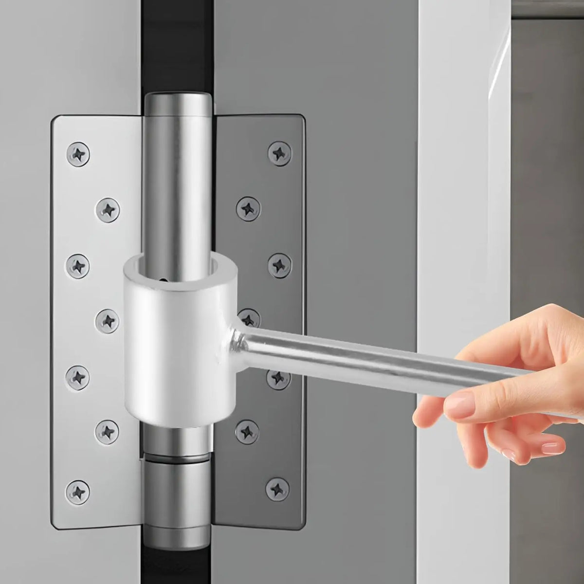 A hand is using a silver heavy duty adjustable door hinge wrench to operate a door hinge on a gray door.