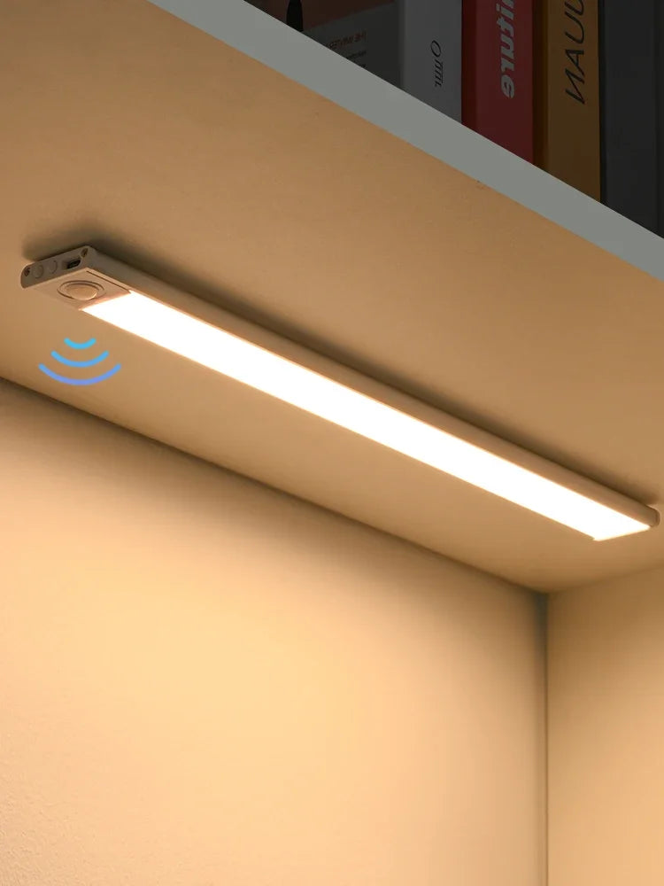 A long, linear LED light bar with a motion sensor is mounted under a cabinet in a beige kitchen setting.