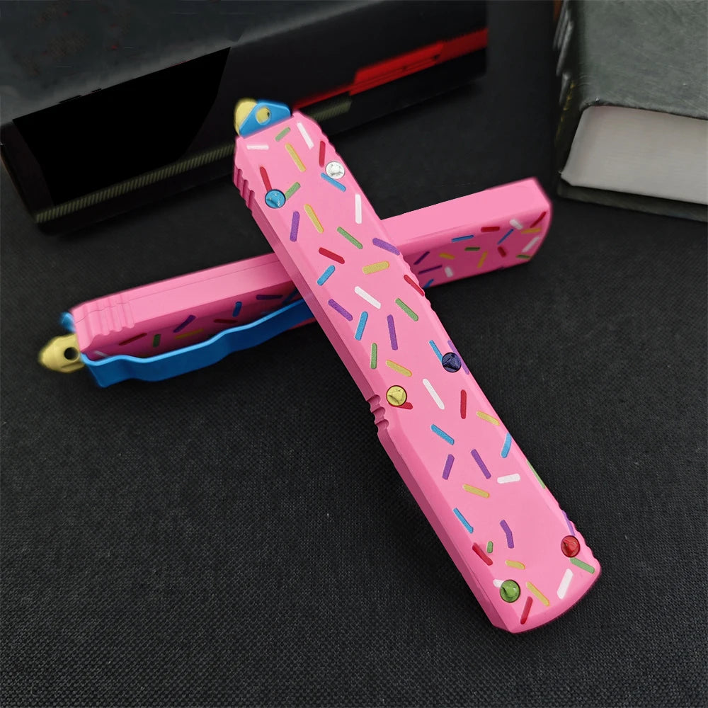 A pink tactical folding knife with a colorful D2 blade and aluminum handle lying on a dark surface.