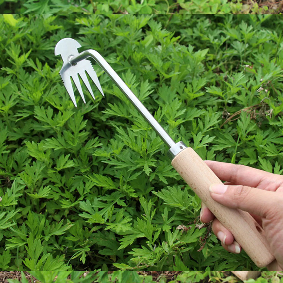 A hand holding a stainless steel fork-like tool for deep root weed removal in green foliage.