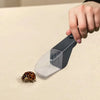 A hand holds a clear plastic pest catcher device with a black handle, positioned over an insect on a light surface.