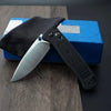A black folding EDC pocket knife with a silver blade is displayed on a blue box and dark wood surface.