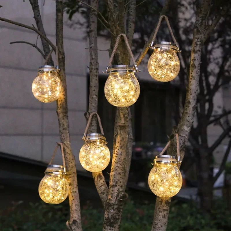 Mehrere leuchtende, knisternde Glas-Solarlaternen hängen von Ästen im Freien.