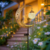 A golden crescent moon light fixture shaped like an owl is mounted on a dark pole in a garden setting.