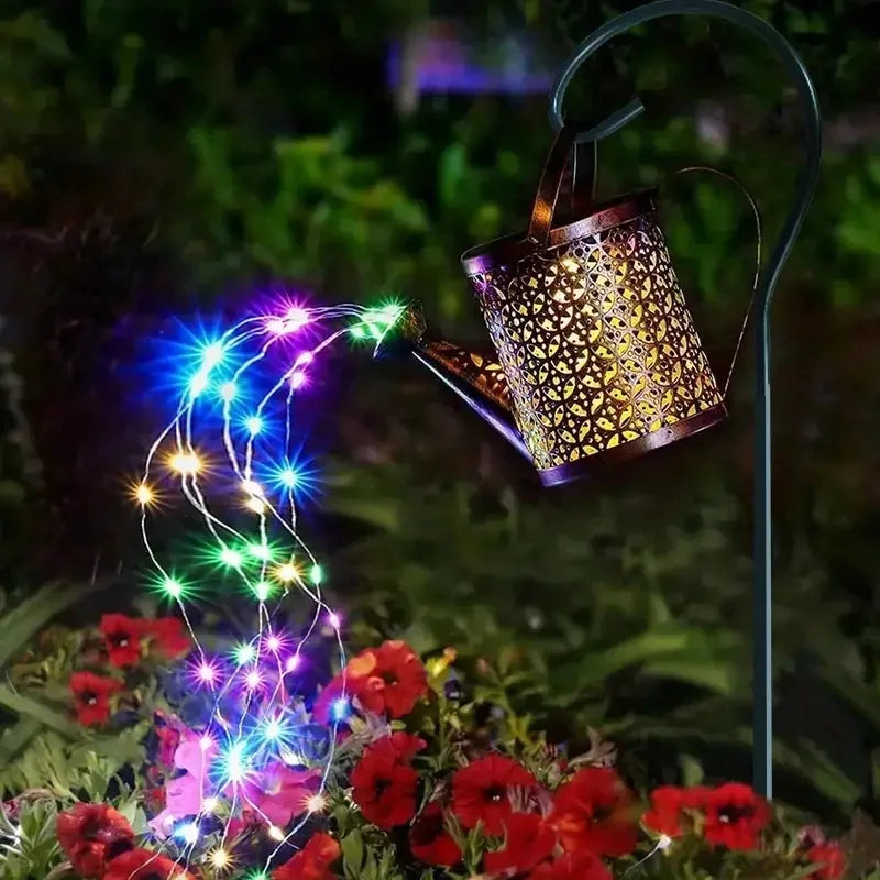 A solar fairy watering can light illuminates a vine-like string of colorful LED lights over red flowers.