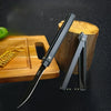 A black outdoor folding knife is displayed next to a wooden block and a yellow knife accessory on a dark surface.