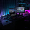 A dark desk setup featuring a laptop, monitor, keyboard, and various electronic components illuminated by purple LED strip li
