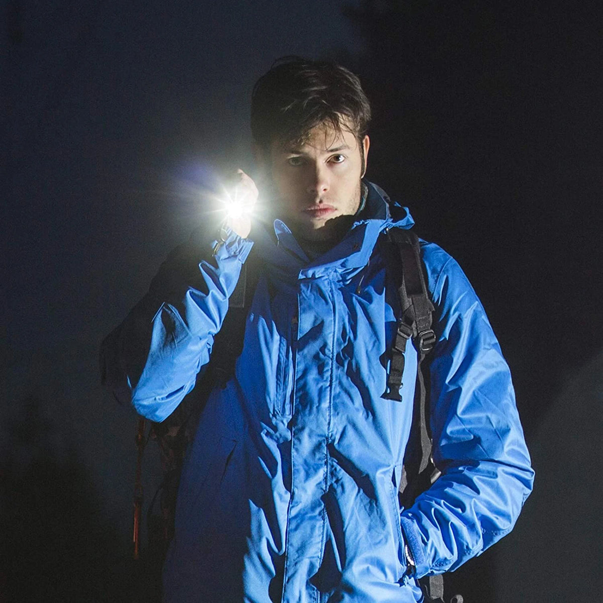 Une personne tient une veste bleue et éclaire son visage avec une lampe torche LED vive sur fond sombre.