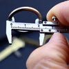 A hand holds a precision metal jewelry measuring ruler and caliper keychain against a dark background.