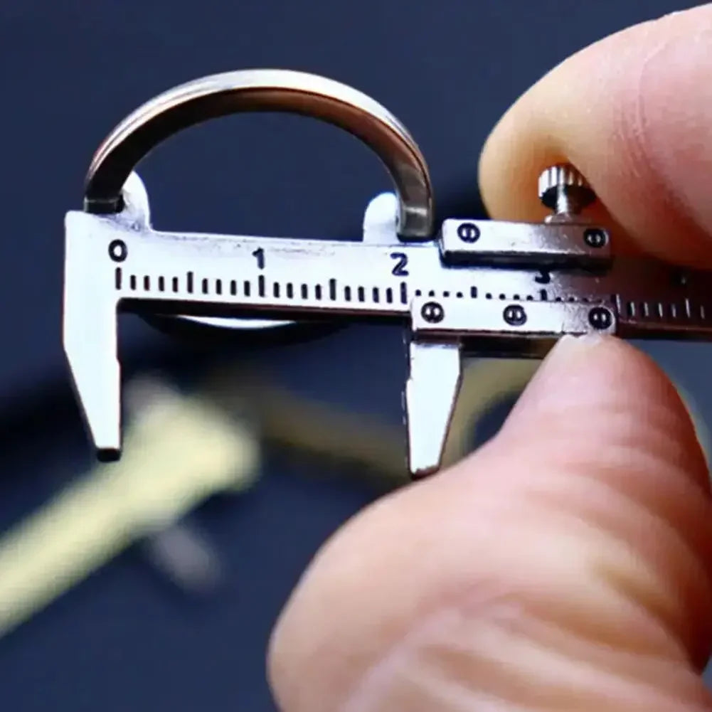 A hand holds a precision metal jewelry measuring ruler and caliper keychain against a dark background.