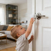 A toddler reaches to operate a no-drill snap-on door lock on a white interior door.