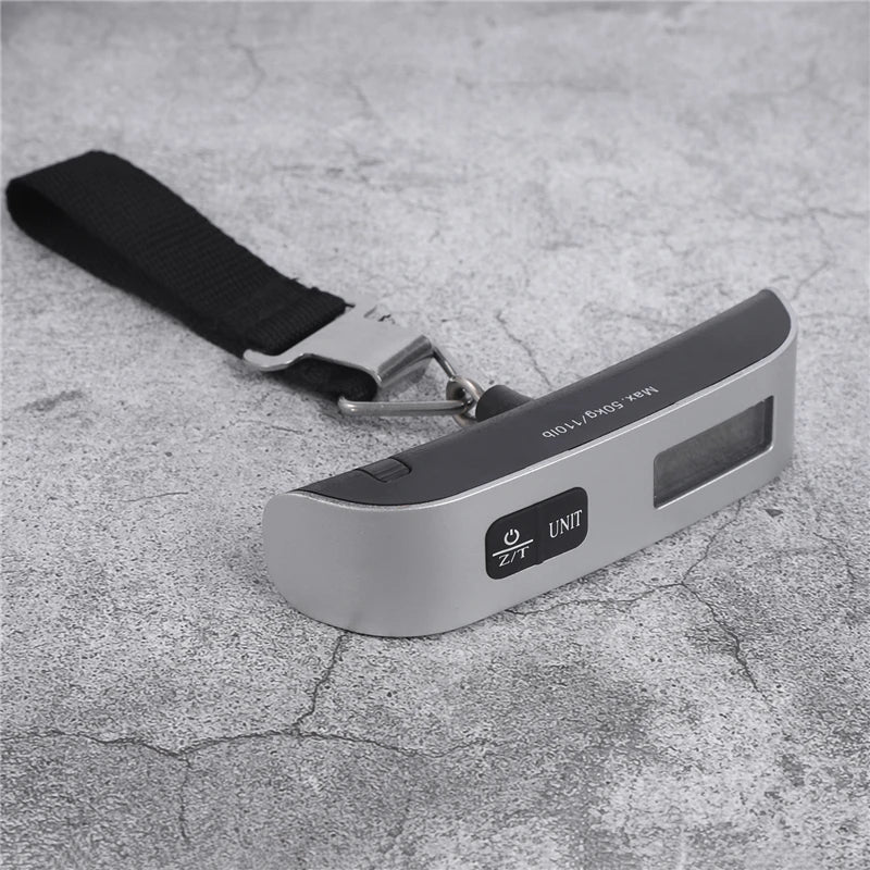 A silver digital weighing scale with a black strap is shown resting on a textured gray surface.
