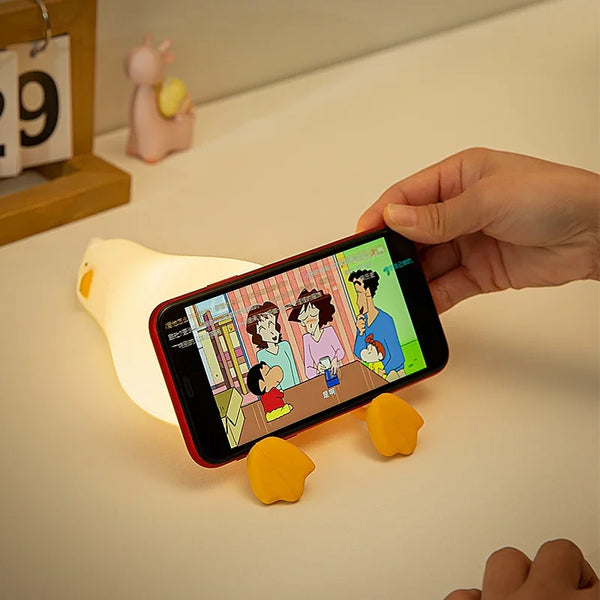 A hand holds a smartphone displaying a cartoon, with silicone duck night lights nearby.