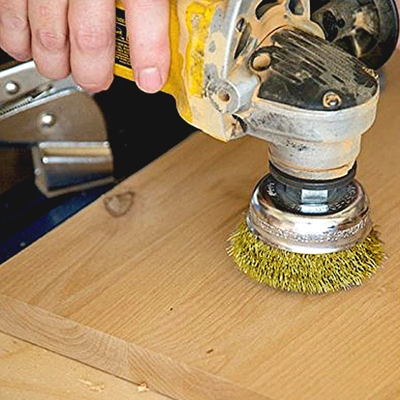A hand is using a yellow and silver wire brush with a green bristles to clean a wooden surface.