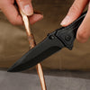 A hand holds an outdoor folding utility knife with a black blade and textured handle, positioned near a dry stick.