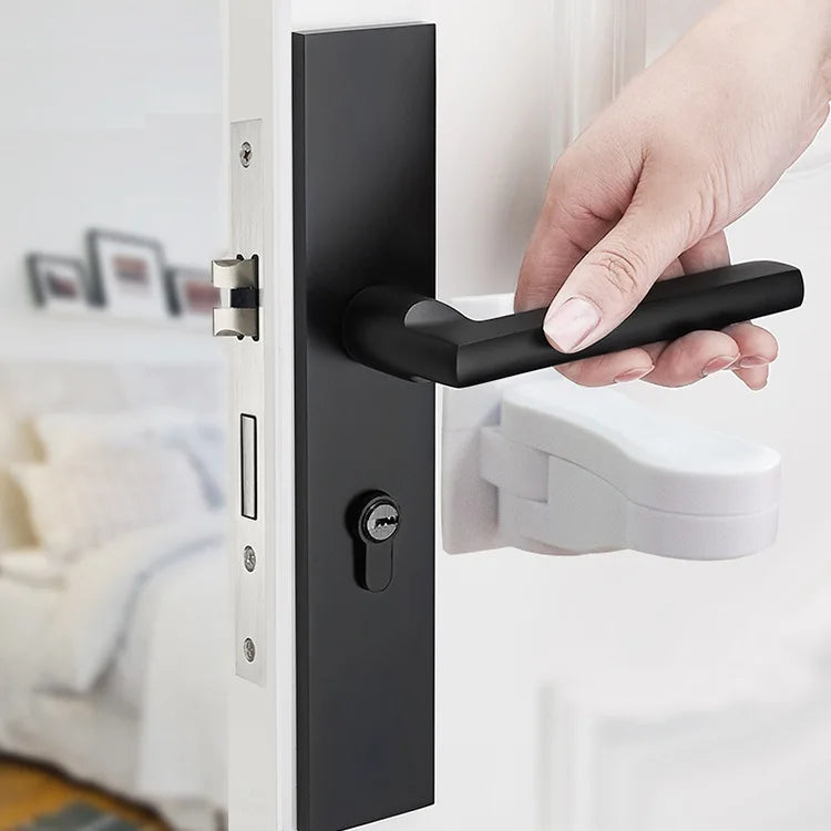 A hand is operating a black universal adhesive door latch on a white door frame for child safety.