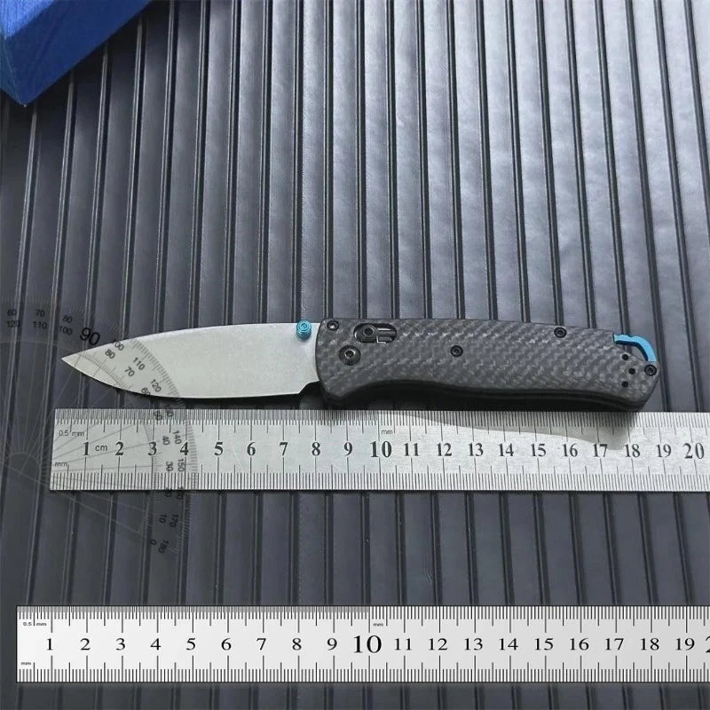 A carbon fiber folding knife blade and handle are shown resting on a ruler against a dark gray background.
