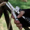 A hand holds a compact multi-tool pocket knife set with silver blades and black grip, resting near a wooden branch.