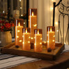 مجموعة شموع عمودية برتقالية متوهجة معروضة على صينية خشبية مع نباتات مزخرفة في الخلفية.