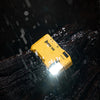 A yellow 400 Lumen rechargeable EDC flashlight is illuminated, shining light through falling rain onto a dark, textured surfa