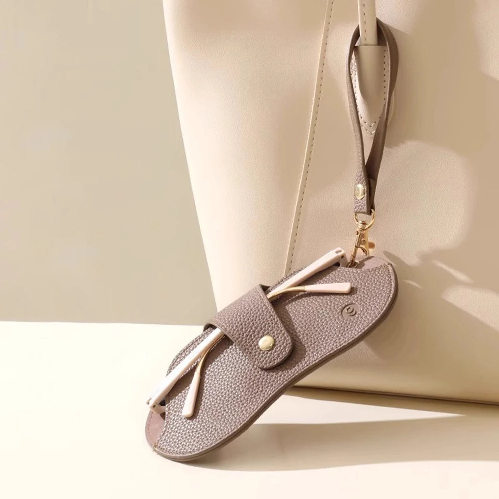 A light taupe Lychee pattern PU leather eyeglasses case with a gold-tone chain strap resting on a cream colored bag.
