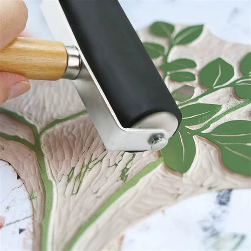 A close-up shot of a professional rubber roller printing tool with a wooden handle and silver metal frame resting on a green 