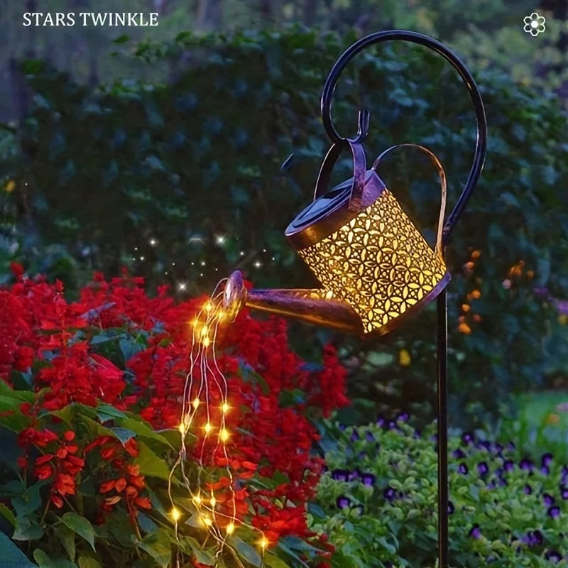 A brass watering can light with glowing fairy lights is shown in a garden setting.