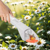 A hand holds a white handheld insect trap with a butterfly inside, set against a background of white and yellow flowers.