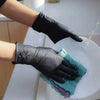 A hand wearing a black disposable nitrile glove is washing a white plate with a green cleaning cloth.